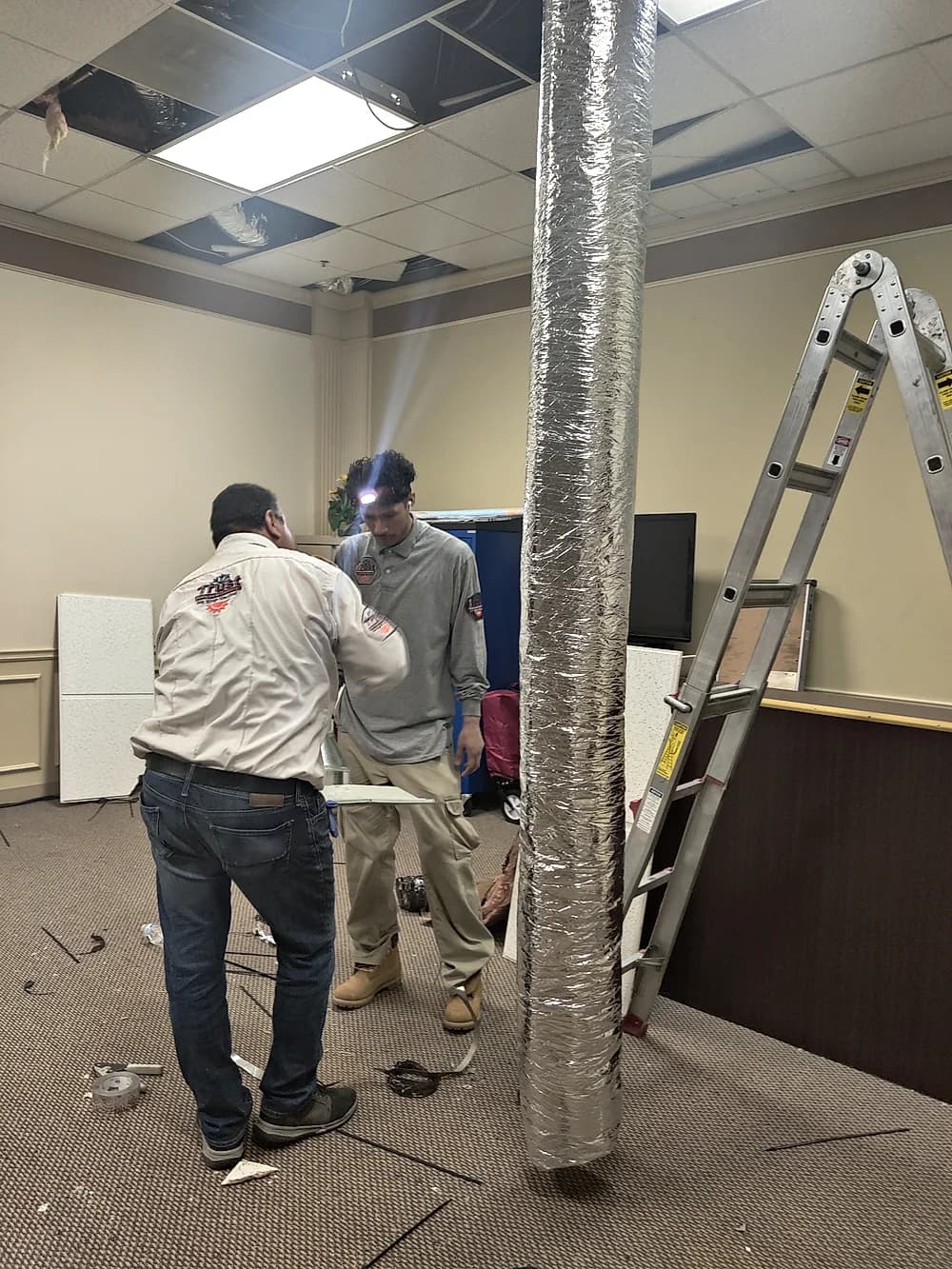 Two Trust Heating and Air Conditioning technicians running insulated ductwork inside a commercial office ceiling